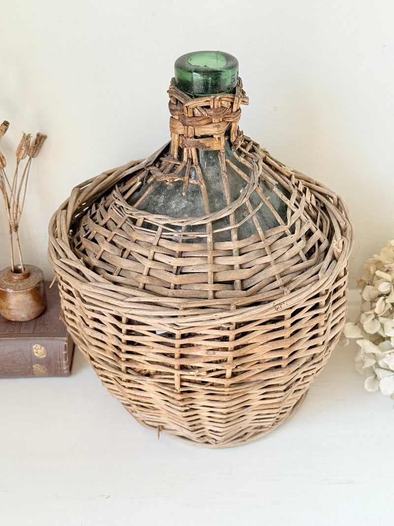antique French demi john olive oil jar in wicker basket with green glass bottle and painted metal label, displayed on white surface