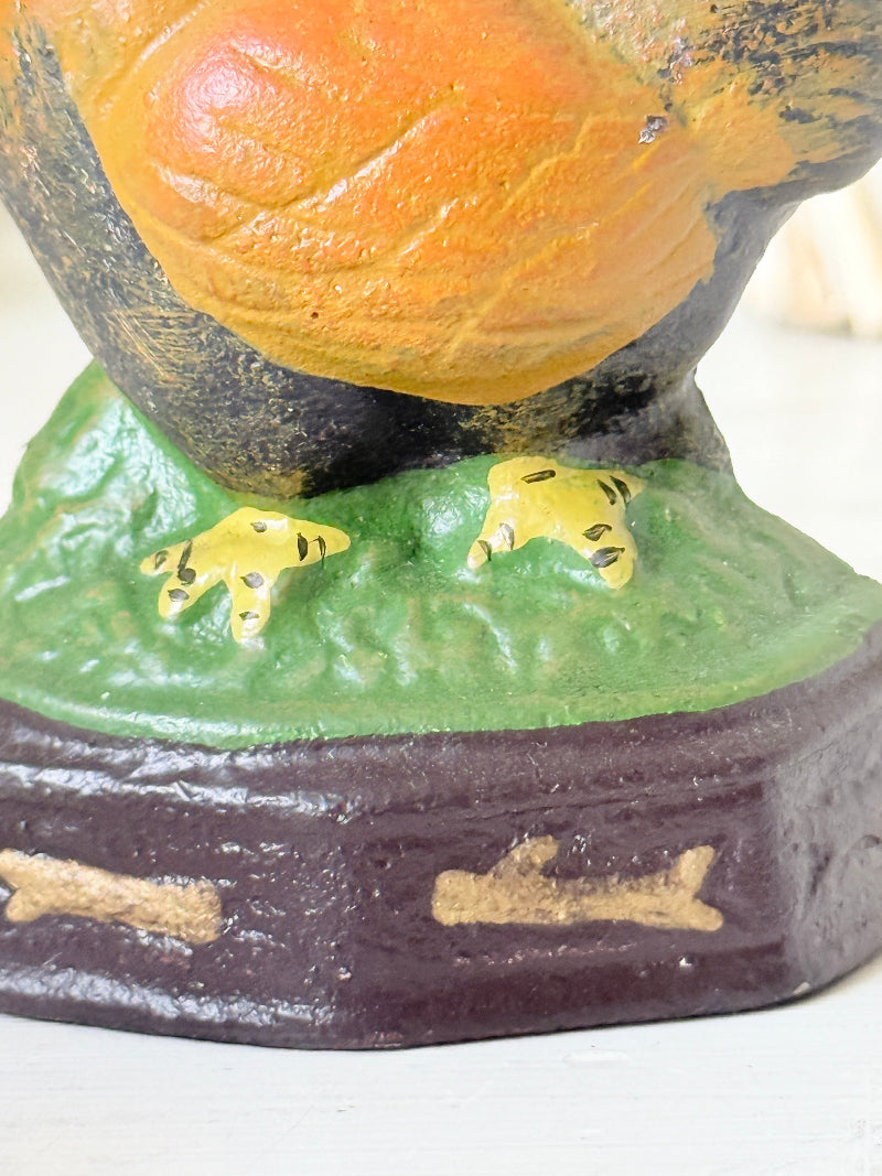 Close-up of a decorative bird figurine with a large orange body, dark head and legs, green textured base, and small yellow feet, shown against a light neutral background.