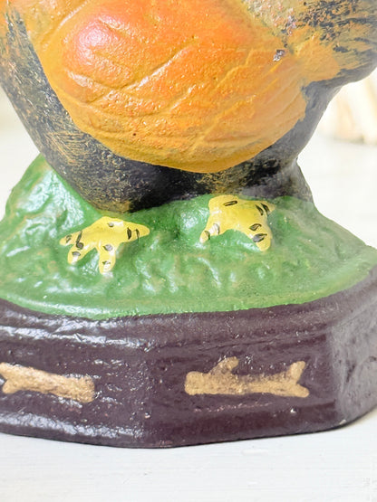 Close-up of a decorative bird figurine with a large orange body, dark head and legs, green textured base, and small yellow feet, shown against a light neutral background.