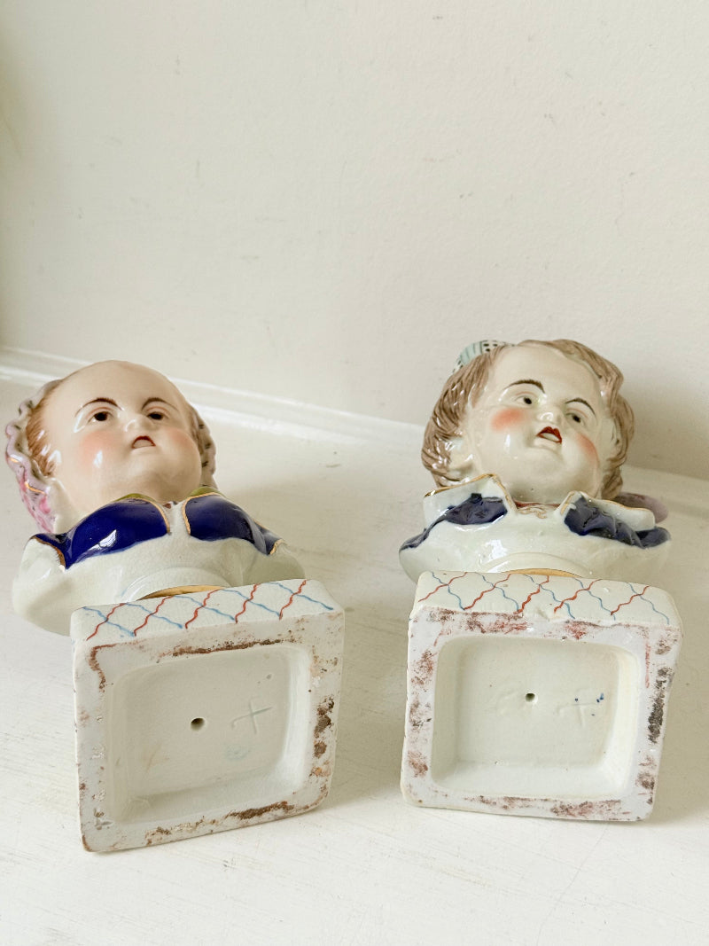 Hand painted Victorian Staffordshire ceramic children busts on white surface, photographed beside dried stems.