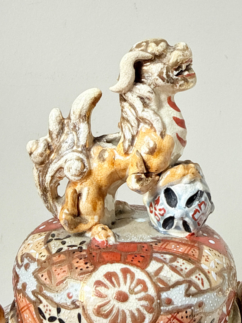 Close-up of a decorative ceramic object featuring a stylized dog or fox figurine in tan, cream, and red glaze, perched on top of a patterned round vessel with floral motifs.
