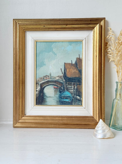 A framed oil painting of a Venetian canal scene with a gondola, displayed in a gold frame on a white surface next to a glass vase with dried grass and a seashell.