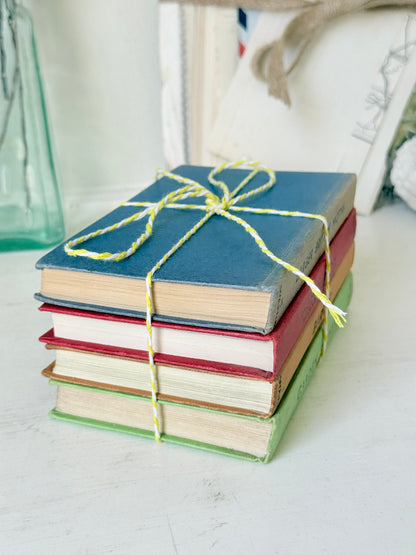 A stack of four vintage hardcover books tied with yellow-and-white twine, set on a white tabletop with a softly blurred background.
