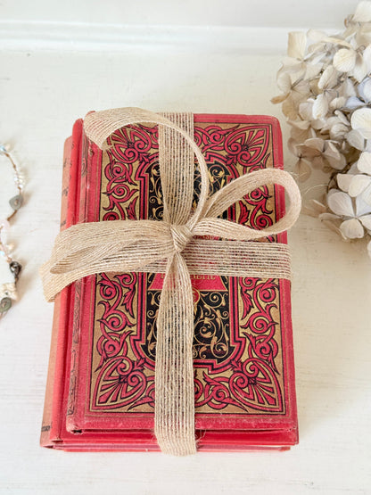 A stack of vintage red hardcover books with ornate gold and black filigree patterns, wrapped in a natural burlap ribbon and tied in a bow, displayed on a white wooden surface.
