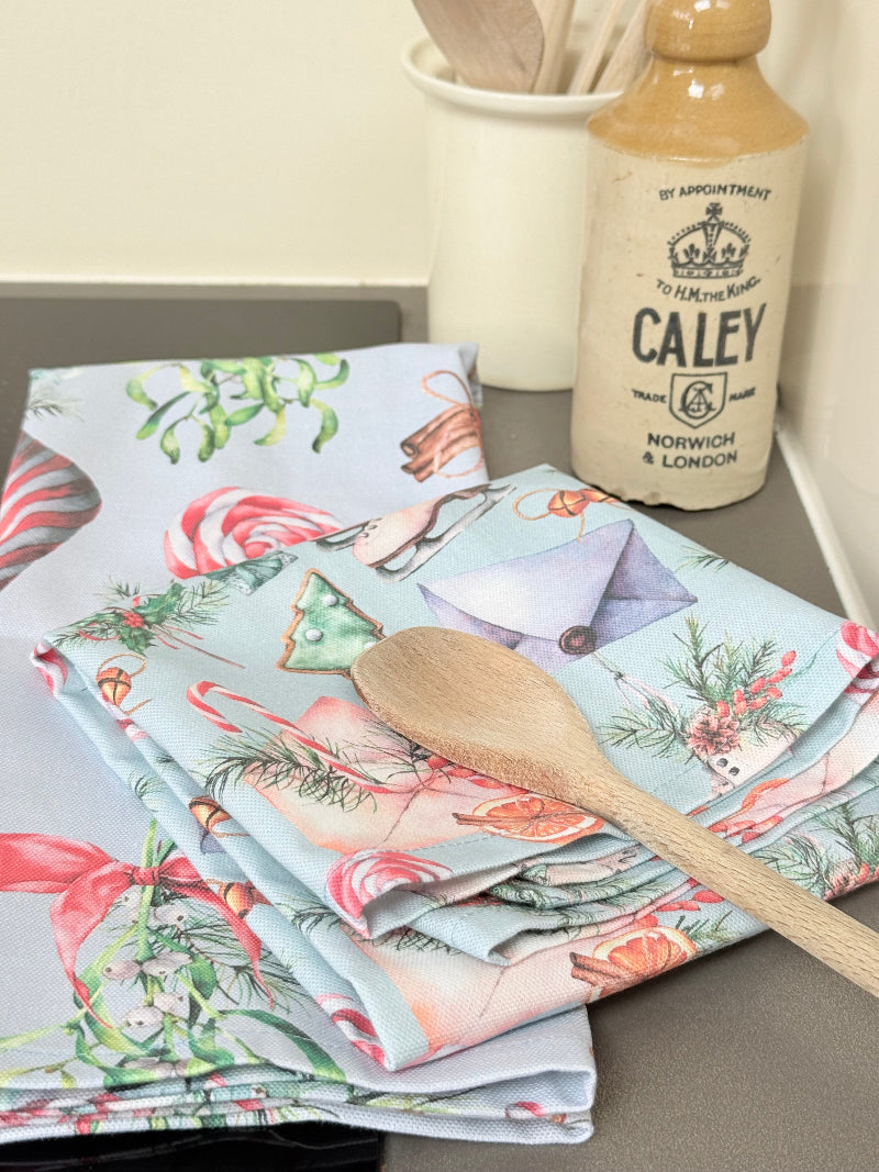 Pair of pastel blue festive tea towels with candy cane and ribbon print, folded, photographed on a kitchen worktop