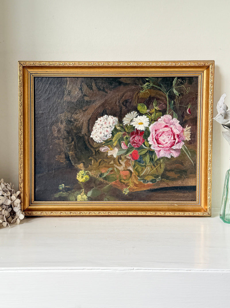 A framed still life oil painting featuring a vibrant bouquet of pink, white, and red flowers in a vase, set against a dark brown background. The painting is housed in an ornate gold frame and displayed on a white shelf.