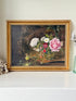 A framed still life oil painting featuring a vibrant bouquet of pink, white, and red flowers in a vase, set against a dark brown background. The painting is housed in an ornate gold frame and displayed on a white shelf.