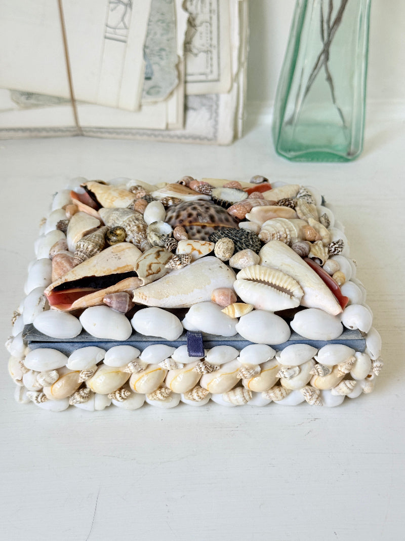 A square decorative box covered with white and beige pebbles and shells, placed on a white surface with a blurred green glass vase and vintage papers in the background.