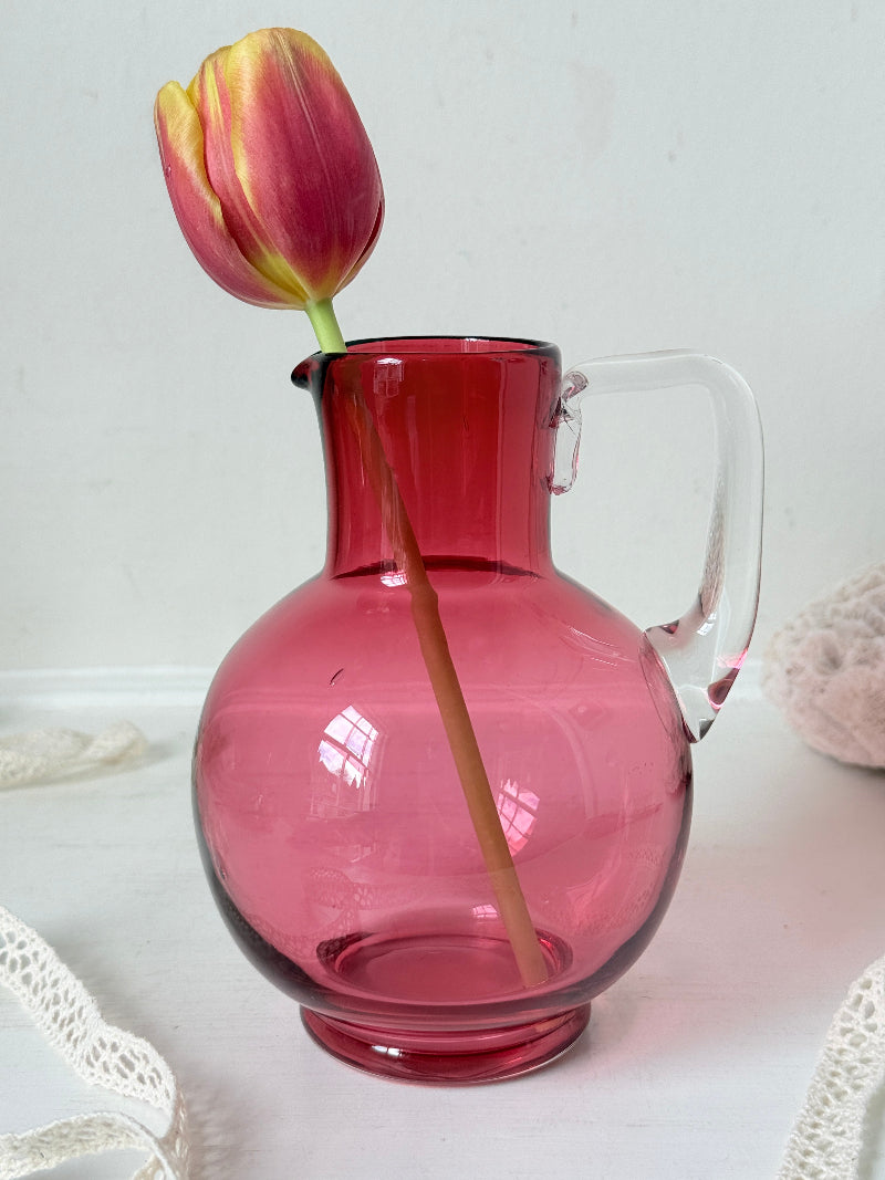 Vintage cranberry glass jug with clear handle photographed on a white surface, bulbous pink body with flared pouring rim