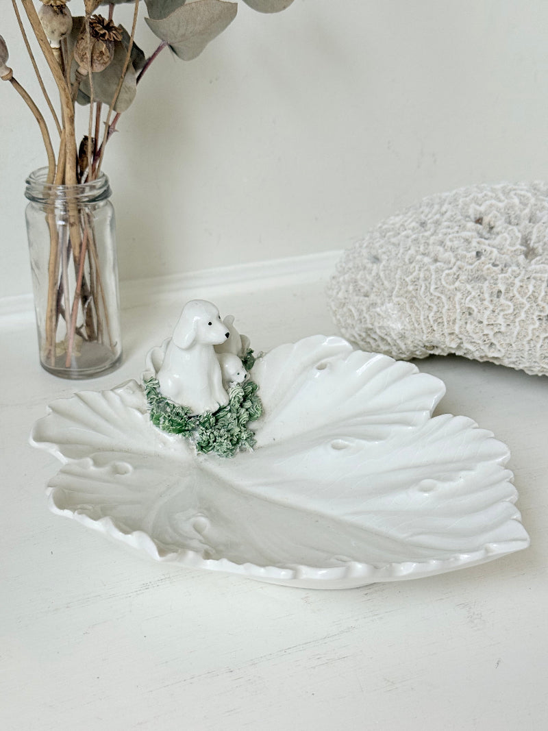 Italian ceramic dish featuring two white dogs on a sculpted leaf base, photographed on a white surface.