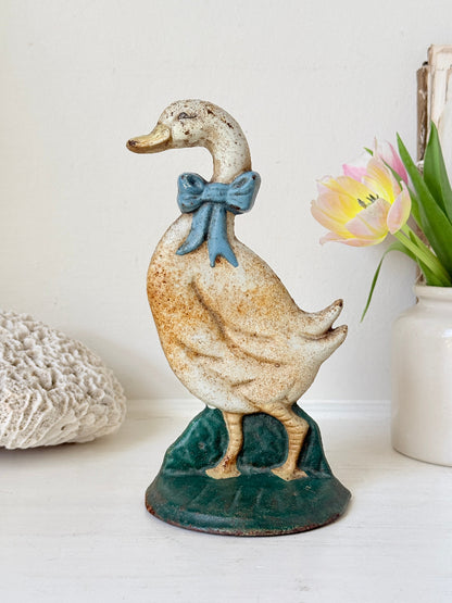 A vintage-style ceramic duck figurine with a speckled cream and brown body, a blue bow, and a dark green base, displayed on a white surface with a vase of tulips in the background.