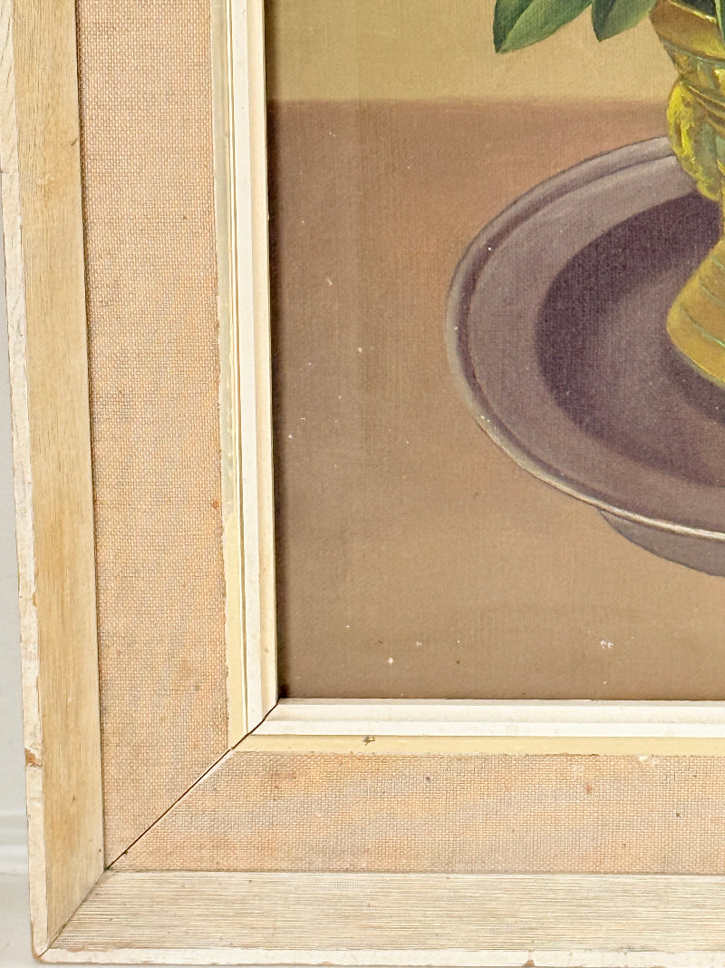 Close-up detail of a framed vintage painting showing a light wood frame, canvas backing, and part of a still life illustration with a green plant and yellow glass vase.