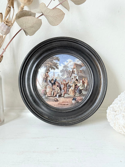 Antique Prattware pot lid in black round frame with rural village scene on white surface.