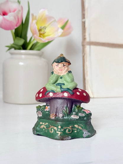 A small hand-painted fairy figurine sitting on a red and white polka-dotted mushroom cap, displayed on a white surface with a blurred white vase of pink tulips in the background.