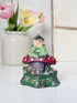 A small hand-painted fairy figurine sitting on a red and white polka-dotted mushroom cap, displayed on a white surface with a blurred white vase of pink tulips in the background.