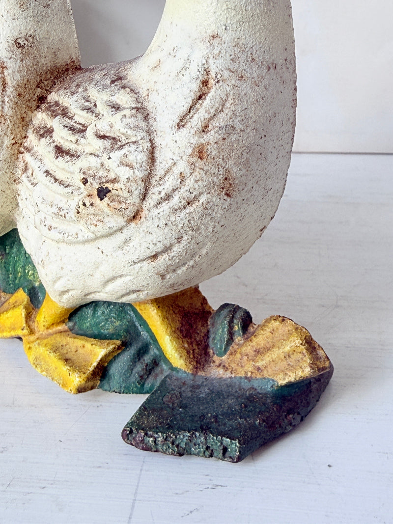 A close-up of a decorative duck figurine or sculpture, showing part of its cream-colored body, yellow beak, and green and gold painted feet on a white surface.