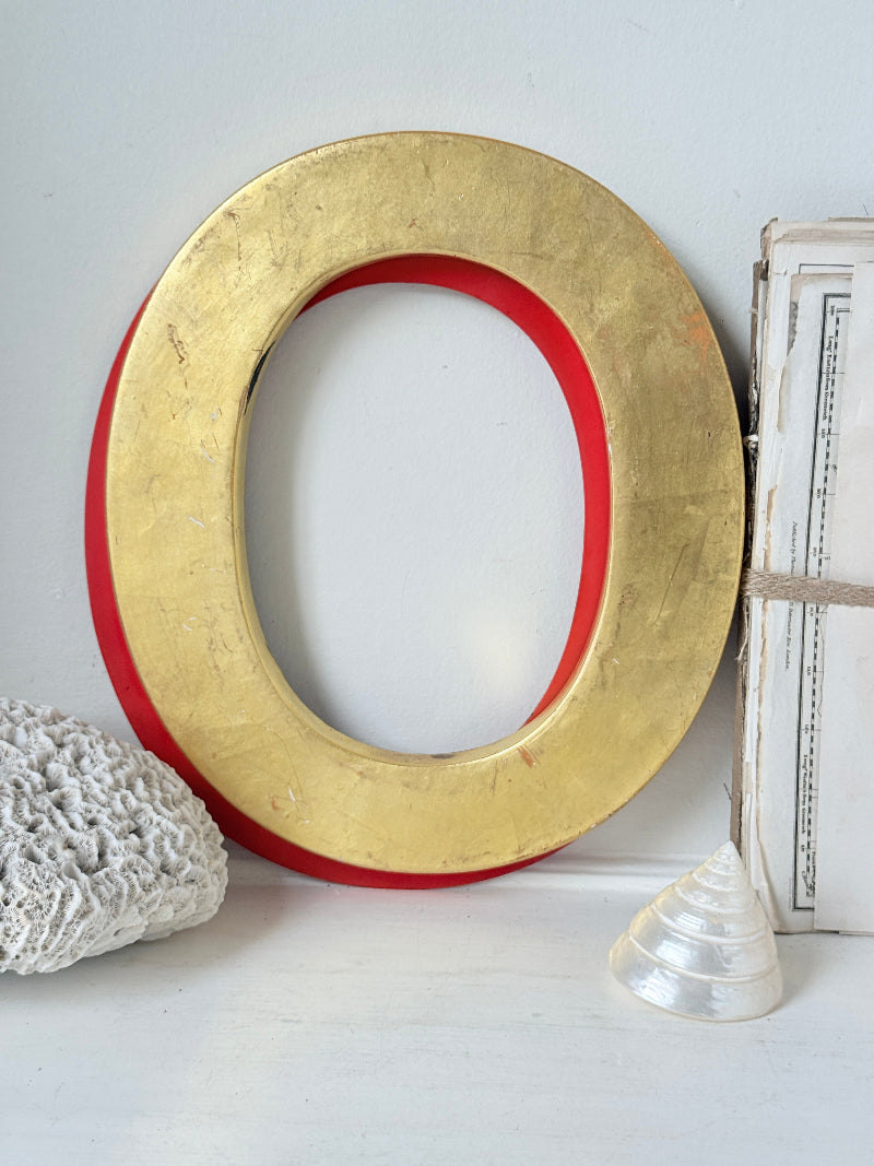 A large decorative letter O with a textured gold outer surface and a bright red inner ring, styled on a white surface with a white woven runner, a small white shell, and a white book or box beside it.