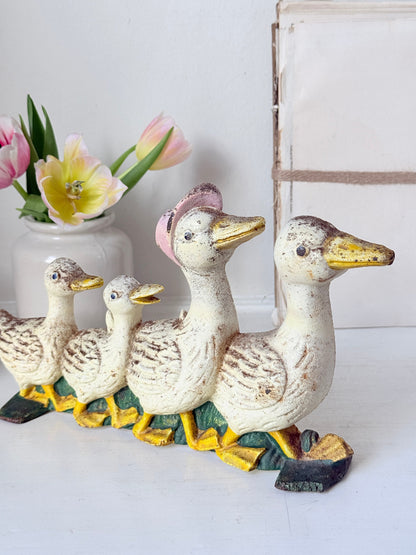 A set of four distressed cast-iron duck figurines, including two adults and two chicks, arranged in a row. The ducks have a cream-colored body with brown speckled detailing, yellow beaks and feet, and a pink feather on the larger duck&