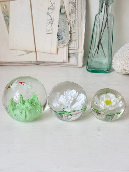 Trio of white flower glass paperweights in graduated sizes with green leaves, photographed on a white surface