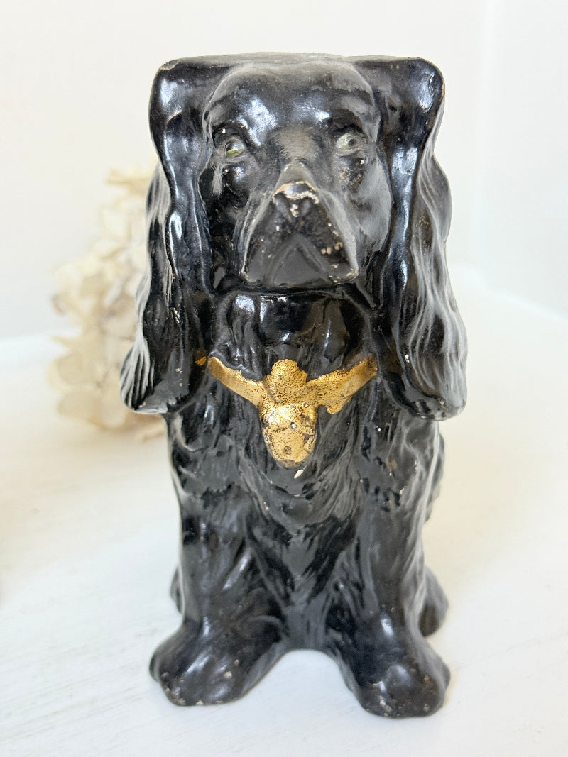 A glossy black ceramic figurine of a long-eared dog, likely a Cocker Spaniel, featuring a metallic gold bird perched on its chest, set against a light background.