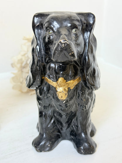 A glossy black ceramic figurine of a long-eared dog, likely a Cocker Spaniel, featuring a metallic gold bird perched on its chest, set against a light background.