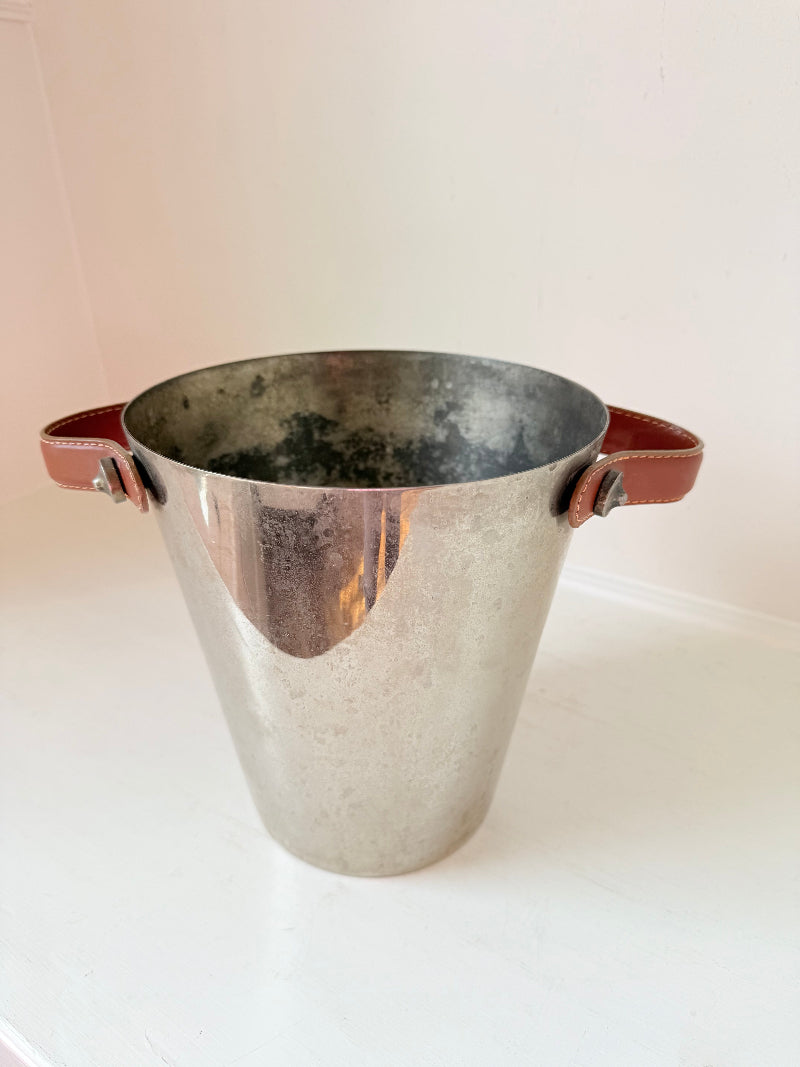 Vintage metal wine cooler with leather handles and tapered form, photographed on white surface.