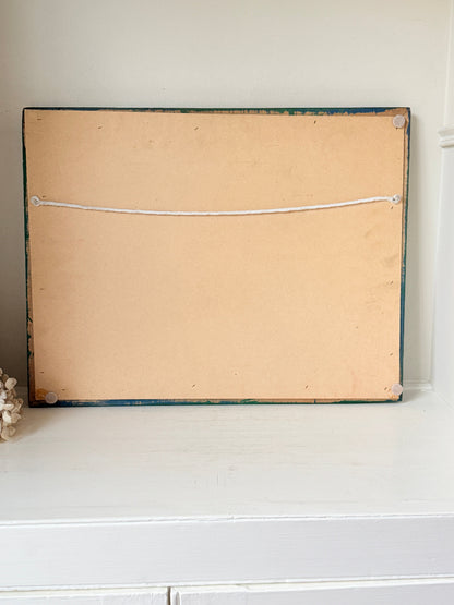 The back of a rectangular framed wall art piece, showing a tan backing board, a thin dark painted frame, and a white hanging cord, displayed on a white shelf against a light wall.