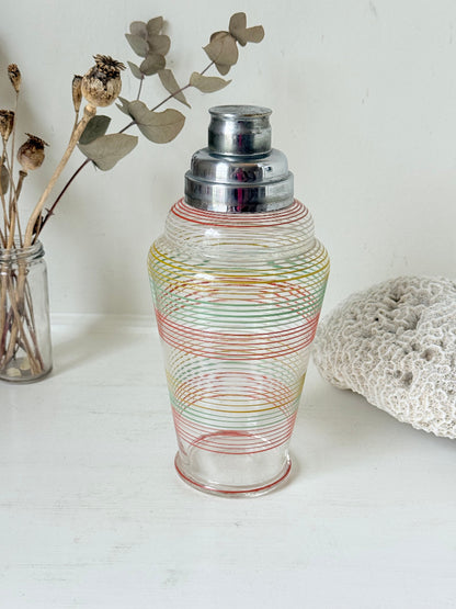 Vintage glass cocktail shaker with multicoloured stripes and silver lid photographed on a white surface.