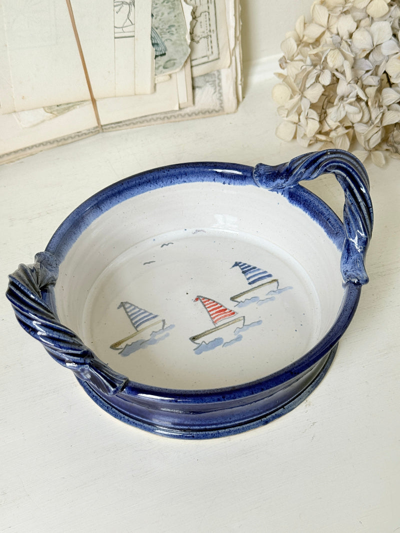 A round ceramic serving dish with twisted rope-style handles, featuring a white interior decorated with three small sailboats in blue, red, and gray, and a dark blue glazed exterior.