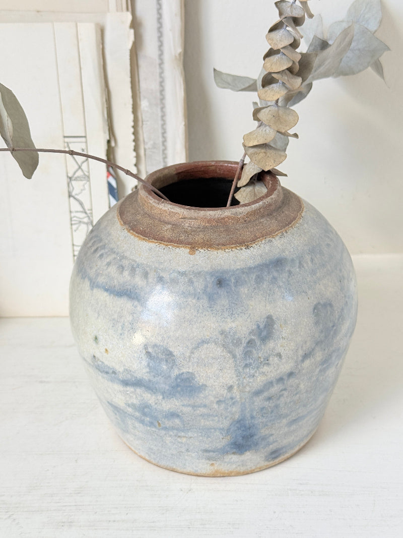 A small round ceramic vase with a blue and white marbled glaze and a rustic brown rim, holding dried eucalyptus stems, set on a white surface against a white background.