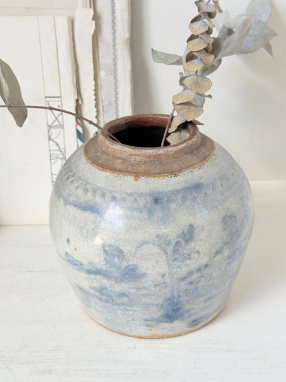 A small round ceramic vase with a blue and white marbled glaze and a rustic brown rim, holding dried eucalyptus stems, set on a white surface against a white background.