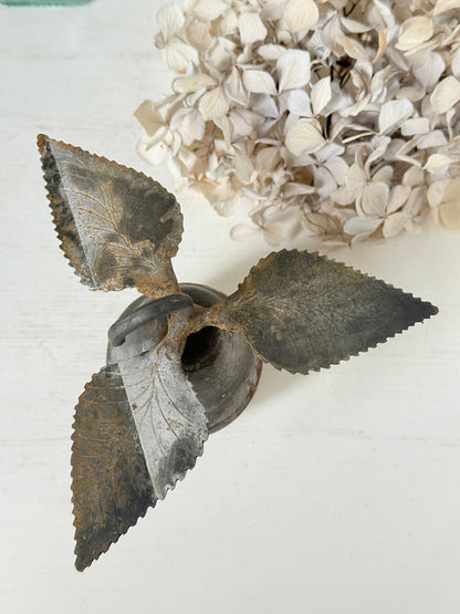 A rustic metal leaf-shaped candle holder with three serrated leaf blades surrounding a central wick, styled on a white surface with dried flowers in the background.