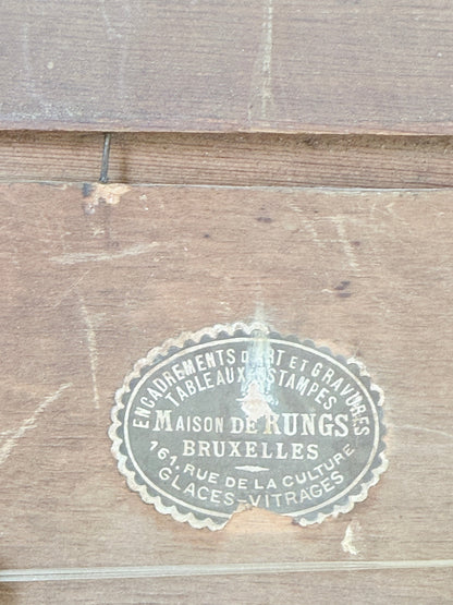 A vintage oval metal stamp for Maison de Rungs Brussels, featuring white text on a weathered silver-gray background, set against a rustic brown wooden plank backdrop.