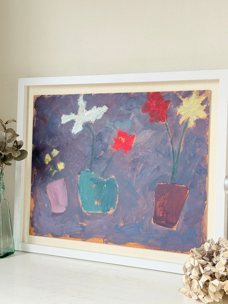 A framed painting of three colorful flowers in pots against a textured purple background, displayed on a white surface with decorative dried flowers and a glass vase nearby.