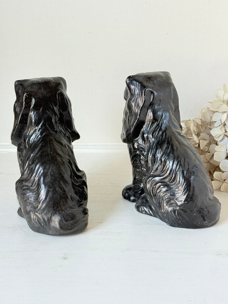 Two glossy black ceramic dog figurines, shown from the back on a white surface against a white wall.