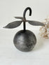 A vintage-style metal hanging ornament shaped like a round engraved sphere topped with a curved hook and leaf-like wings, displayed against a light neutral background with dried flowers at the side.