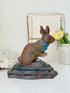 A vintage-style metal rabbit figurine with a textured brown finish and a bright blue bow tie, sitting on a dark base. The figurine is styled on a white surface with a white ceramic vase containing pink tulips and a rustic book in the background.