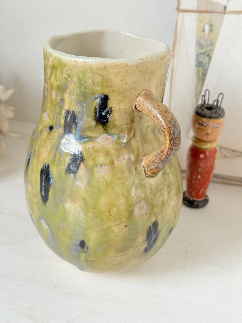 A handmade ceramic pitcher with a rounded body, a curved handle, and a textured glaze in olive green, beige, and black speckles, shown on a white surface with a small toy figure in the background.