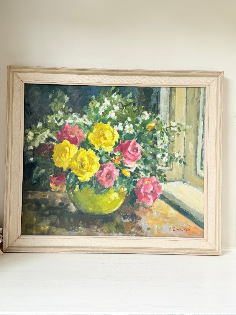 A framed oil painting of a vibrant flower bouquet with yellow and pink roses in a green vase, set against a window background. The painting is housed in a light-colored wooden frame and displayed on a white surface against a neutral beige wall.