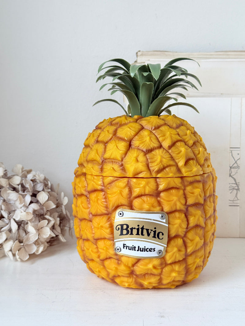 A novelty container shaped like a pineapple, with a textured golden-yellow body, green leafy crown, and a silver label reading &