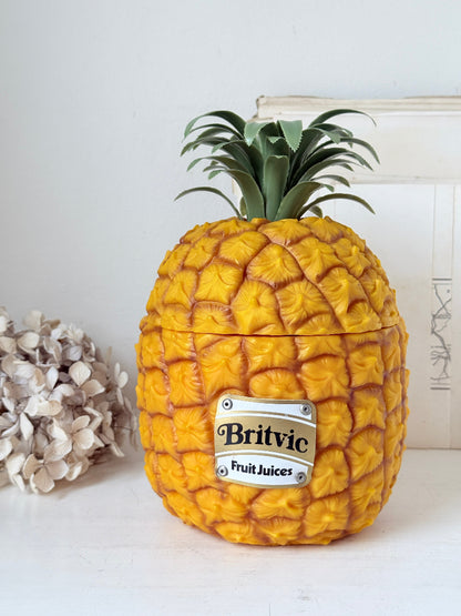 A novelty container shaped like a pineapple, with a textured golden-yellow body, green leafy crown, and a silver label reading &