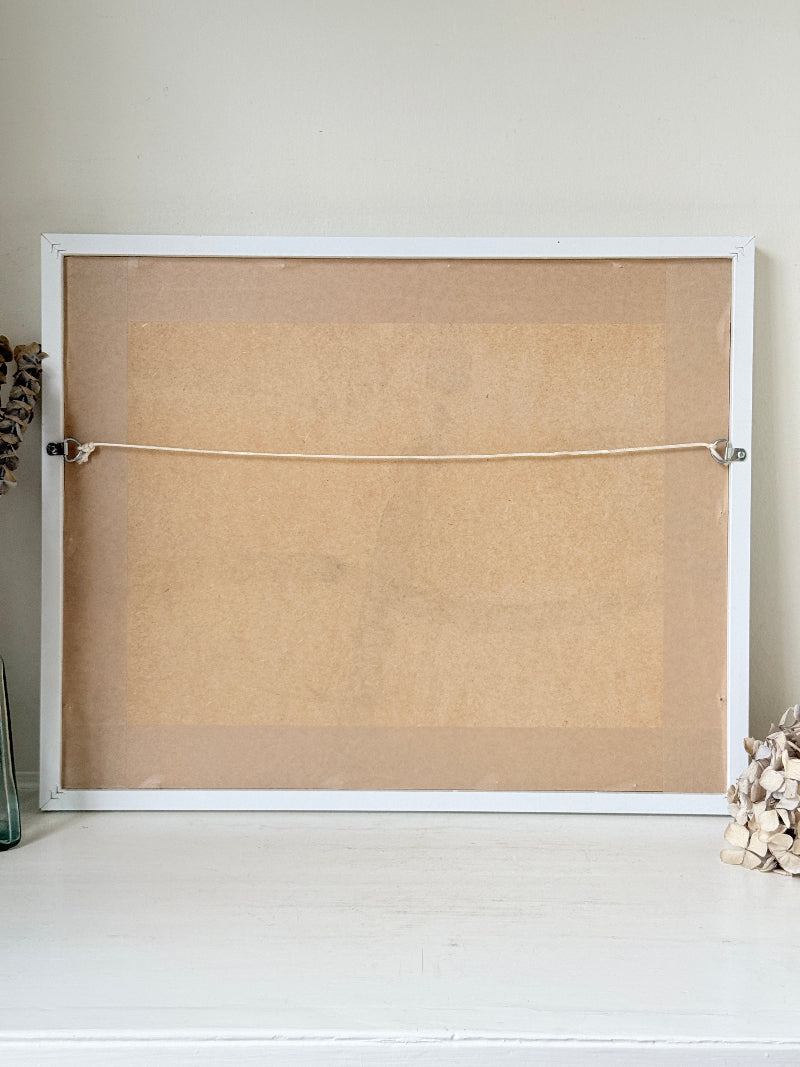 The back of a white-framed cork board or bulletin board, showing the tan cork surface, a horizontal hanging cord, and metal clips, set against a light beige wall and white surface.