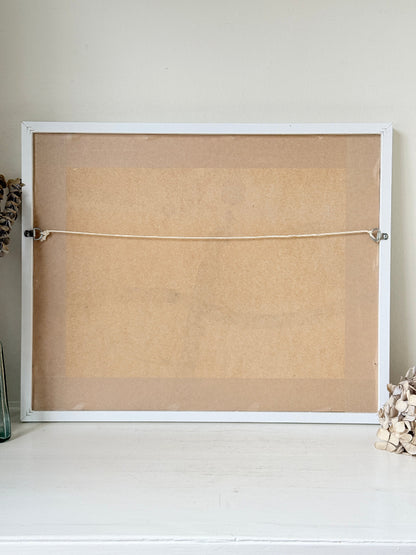 The back of a white-framed cork board or bulletin board, showing the tan cork surface, a horizontal hanging cord, and metal clips, set against a light beige wall and white surface.