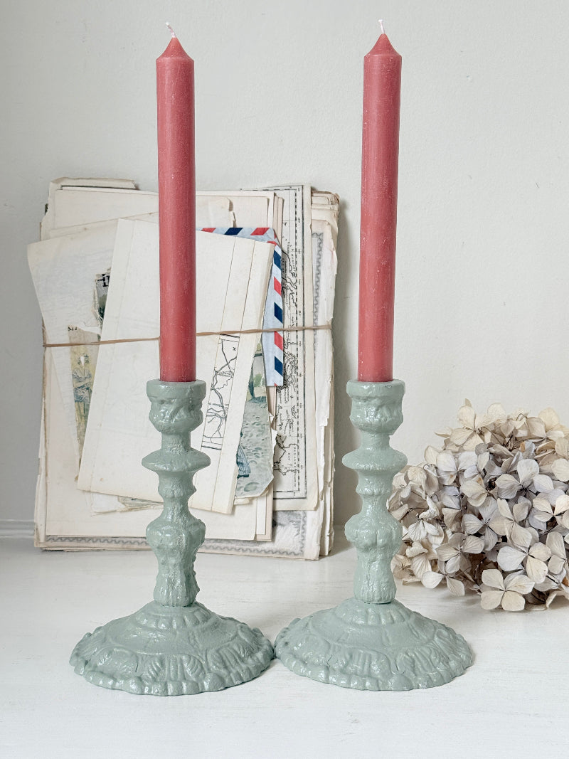 A pair of ornate sage green candlestick holders with tall dusty red taper candles, styled on a white surface with vintage papers and dried flowers in the background.