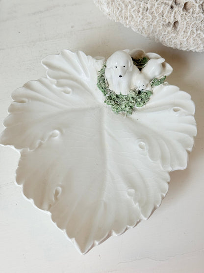 Italian ceramic dish featuring two white dogs on a sculpted leaf base, photographed on a white surface.