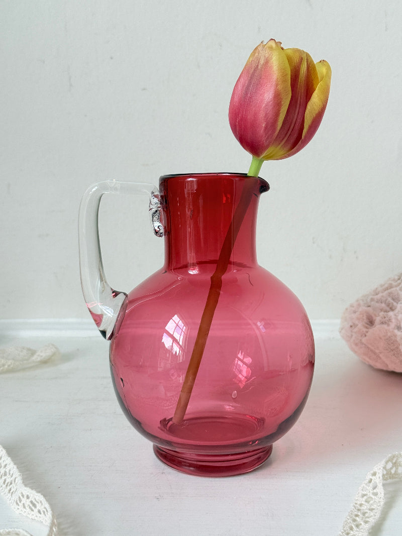 Vintage cranberry glass jug with clear handle photographed on a white surface, bulbous pink body with flared pouring rim