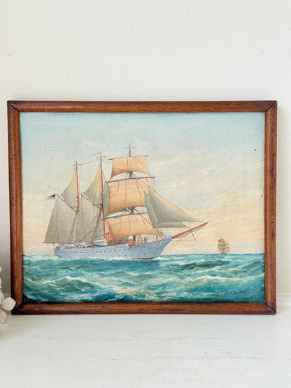 A framed vintage oil painting depicting a white sailing ship with beige sails navigating blue ocean waves under a pale sky, housed in a dark brown wooden frame.