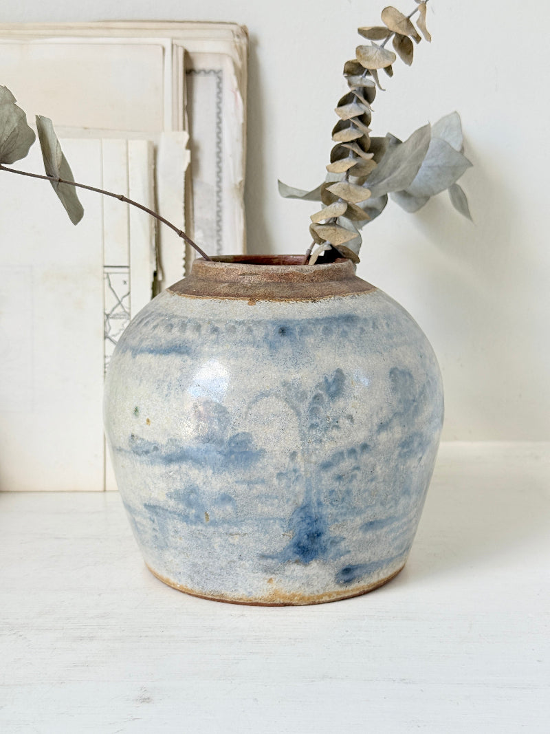 A rustic ceramic vase with a blue and white marbled glaze, holding dried eucalyptus stems, set on a white wooden surface against a white wall.