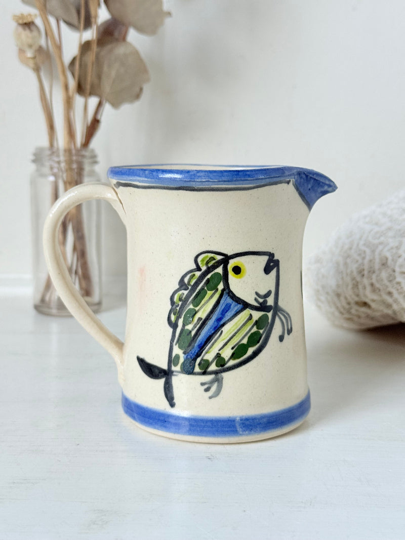 Vintage studio pottery jug with hand painted blue and green fish design on cream clay, photographed on a white surface.