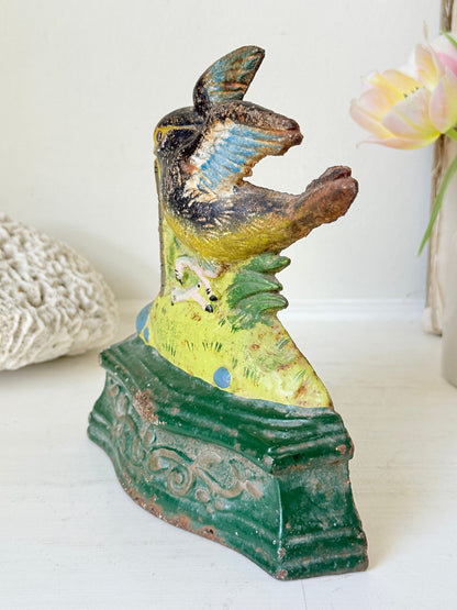 A vintage hand-painted ceramic bird figurine on an ornate green base, displayed on a white surface with soft-focus decorative props in the background.